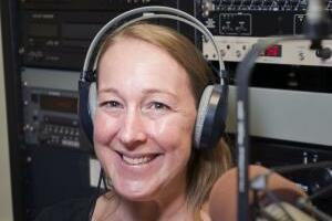 A person smiling with headphones on sitting in front of a microphone