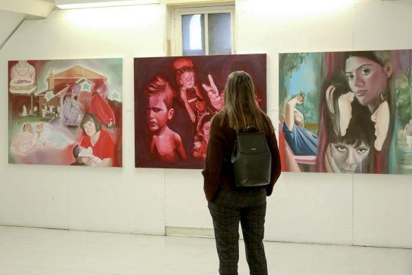 Person standing in a gallery looking at 3 paintings.