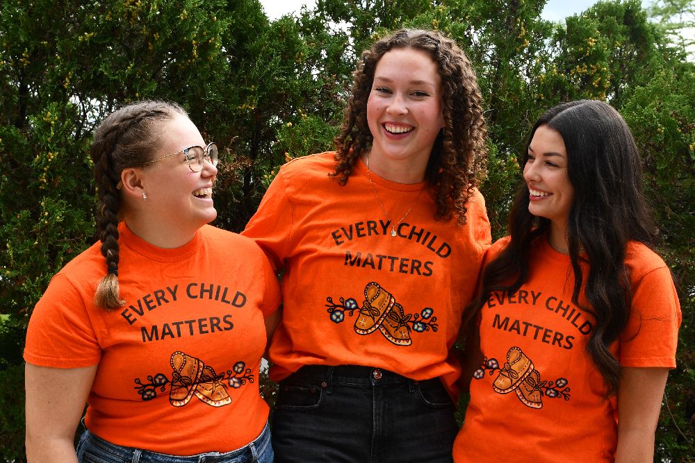 Three people stand wearing the new Orange shirts in front of trees.