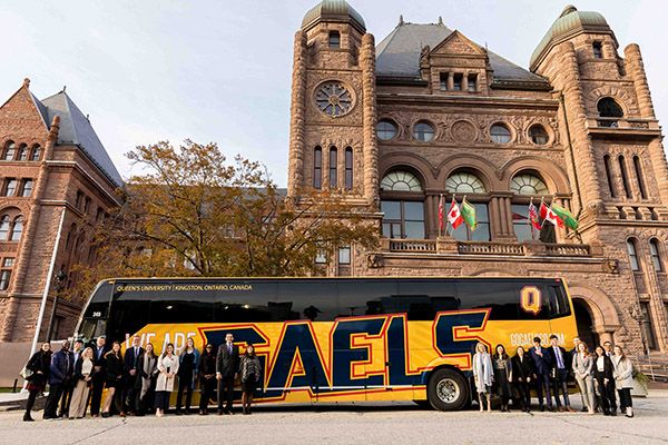 A group of people stand in front of a bus that says "Gaels" on it which stands in front of Queen's Park.