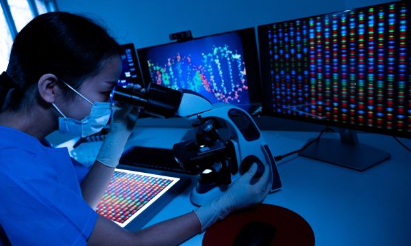Woman wearing a mask looking through a microscope with screens in the background displaying DNA