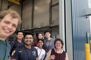 Link to Article: "The BVEX Team Arrives in Timmins". Picture shows seven members of the BVEX team standing in front of the door to a tall building with a large door that is several stories high.