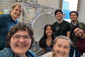 A group of seven people standing in front of a small radio dish mounted on an aluminum gondola frame.  The people and gondola are in a building with a very tall ceiling.