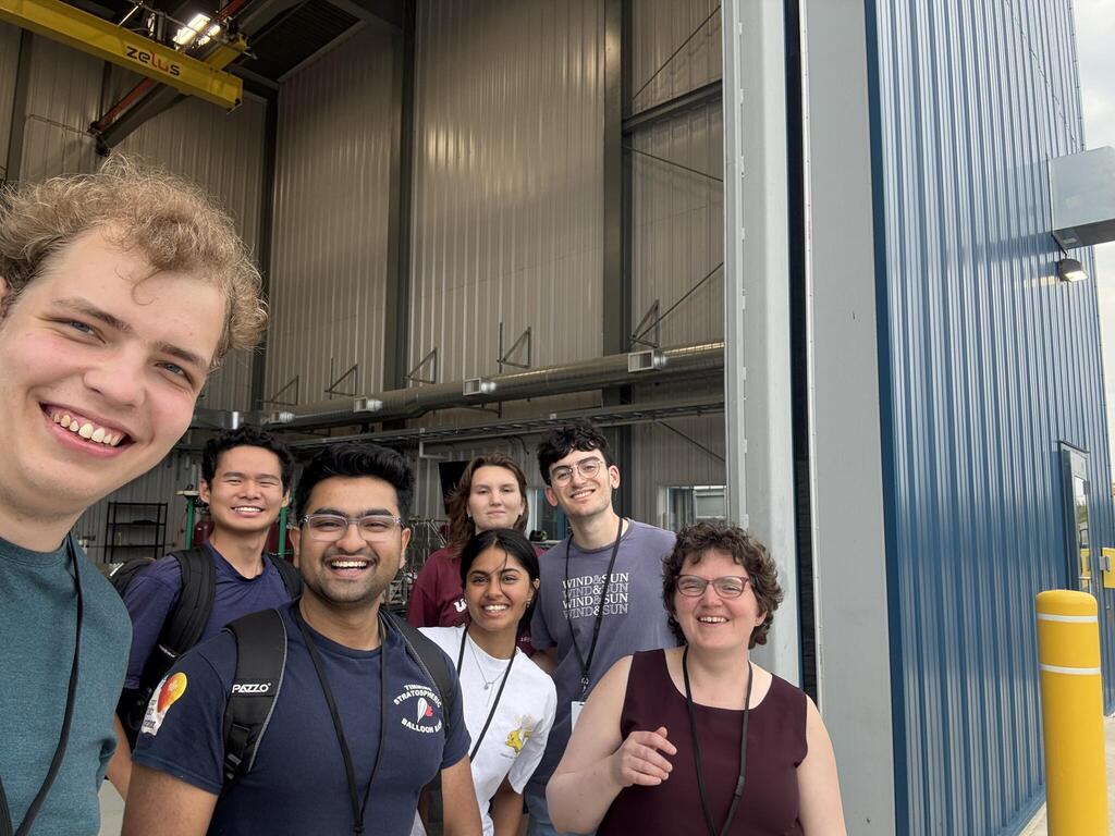 A group of seven people from the BVEX team stands in front of the open door of a building.  The door is several stories high.