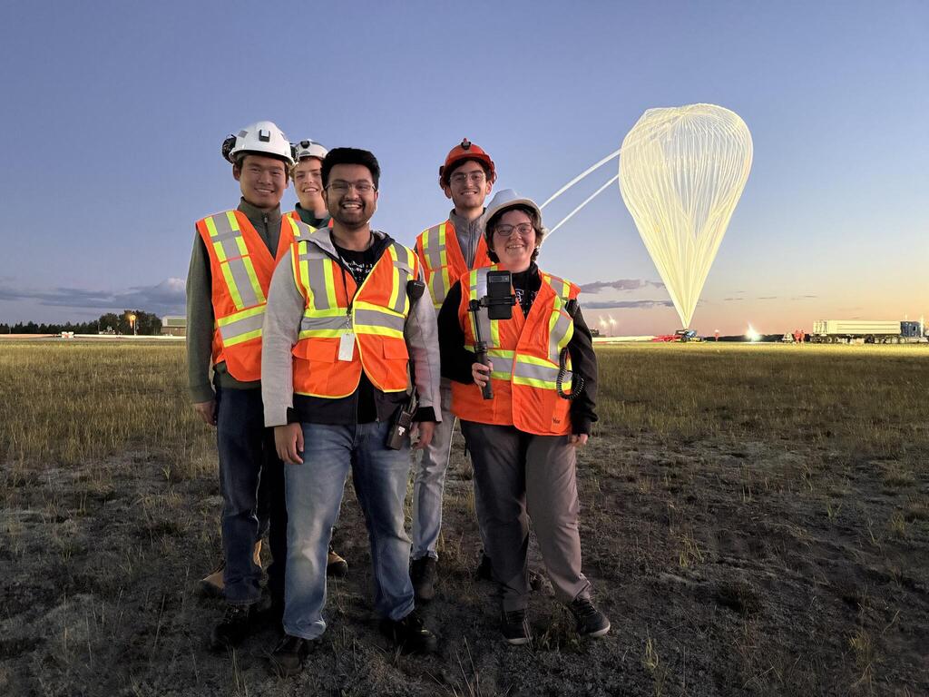 A group of five people wearing safety vests (some with hard hats) standing on an airport tarmac with a large helium balloon in the background.
