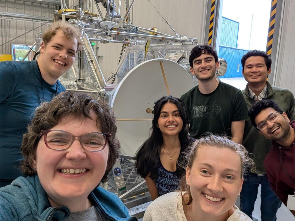A group of seven people standing in front of a radio telescope dish on an aluminum balloon gondola. They are located inside a building with a high ceiling..