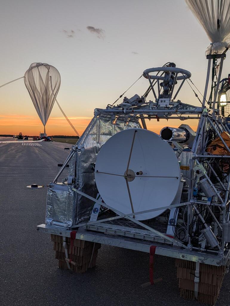 A small radio telescope on a metal structure sits on an airport tarmac at sunset.  In the background a clear plastic large helium balloon can be seen.  Closer behind the telescope in the upper right part of another large helium balloon can be seen.