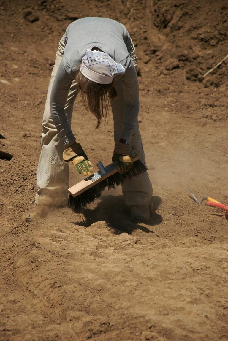 Student using a broom head to carefully sweep away dirt during excavation in Matera.