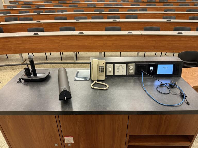 Top down view of the podium. The podium has a set of microphones, a document camera, lecture capture, display cables, and a telephone. The podium is not height adjustable, but it is accessible.
