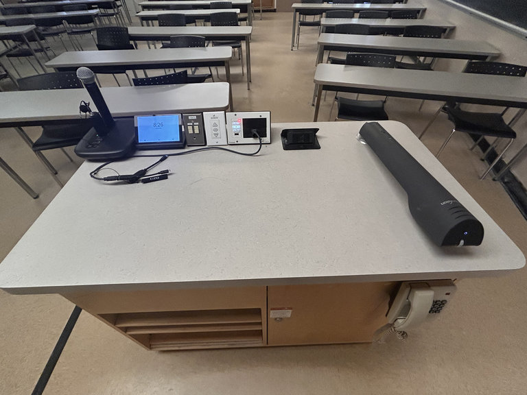 Top-down view of podium. Microphones on the left; touch panel control system at center; document camera on the right; telephone mounted on the side cabinet.
