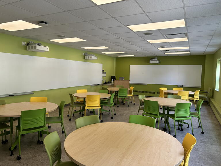 View of the classroom from a student seat near the rear of the room. In front are multiple round tables and chairs. The instructor podium is at the front of the room on the left. Digital projectors on the ceiling, projects onto screens on the front wall and left wall. This classroom is accessible for students.