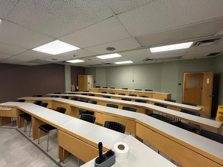 View of the classroom from the instructor’s podium. There are rows of long tables with chairs for students. The row at the back of the classroom is on a raised platform. The entryway door is located at the back of the room, on the left side. This room is 
