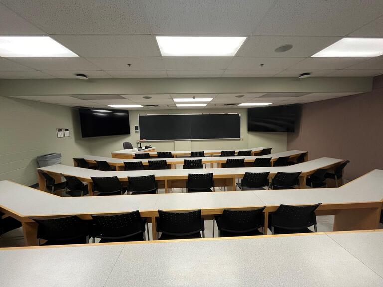 View of the classroom from a student seat near the rear of the room. In front are empty rows of tables and chairs. The instructor podium is at the front of the room on the left. The front of the classroom has a large blackboard in the middle of the front of the room, with two TV's on either side.