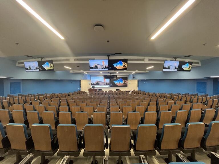 This image depicts the students view when sitting at the rear of the room. There are rows of seating with tablet arms. The room slopes downwards towards the front of the room. There is accessible seating at the front and rear of the room. The instructor’s podium is in the front left.