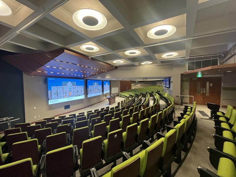 View of the classroom from a student seat at the rear of the room. Instructor’s podium is located at the front of the room. A whiteboard is located on the right wall, close to the instructor’s podium. There are three video walls at the front of the room. 