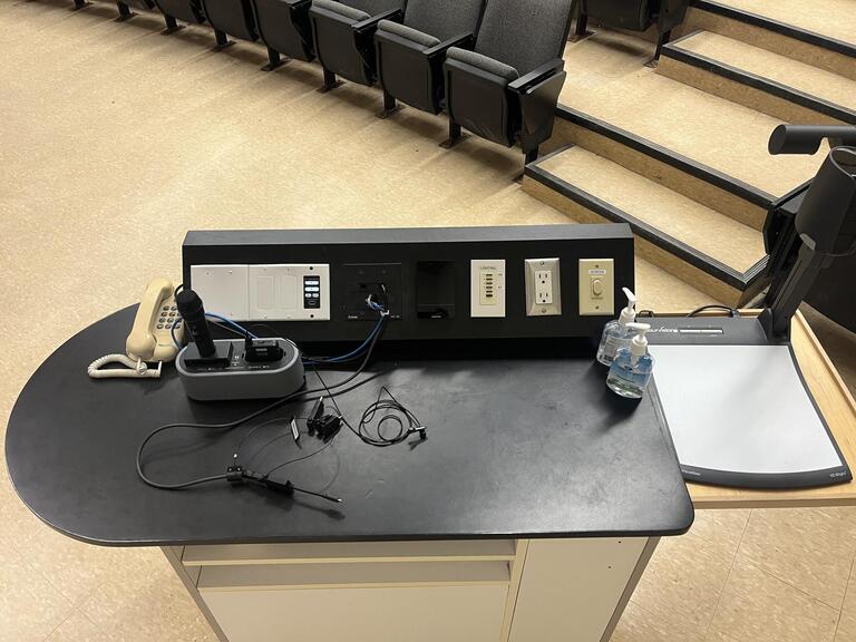 Top down view of the podium. The podium has a telephone, a set of microphones, a lecture capture, and a set of display cables.