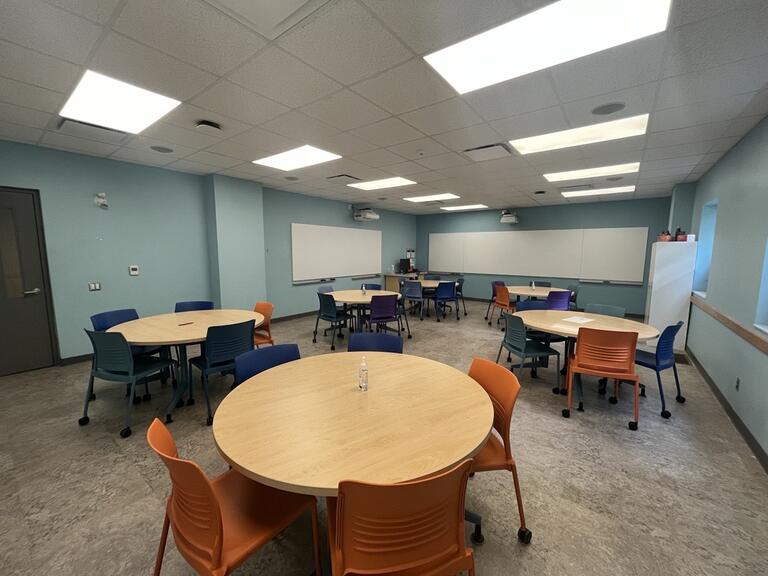 View of the classroom from a student seat near the rear of the room. In front are multiple round tables with chairs. The instructor podium is at the front of the room on the left. Two projectors project onto screens that are located across the front wall, as well as the left wall (from the perspective of a student). This classroom is accessible for students.