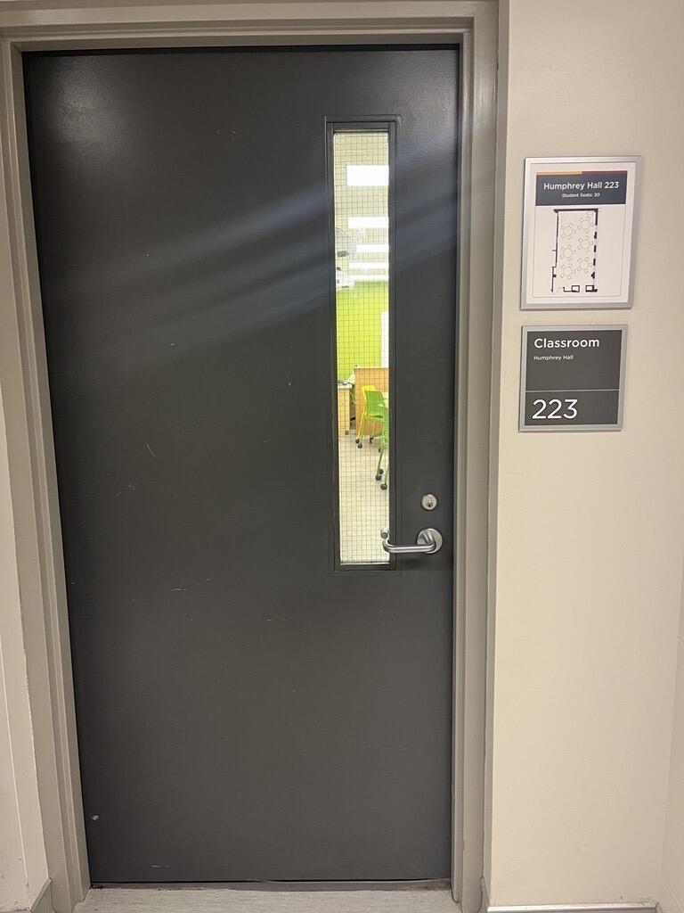 Image of the door to enter Humphrey Hall room 223. The door is grey with a lever on the right side.