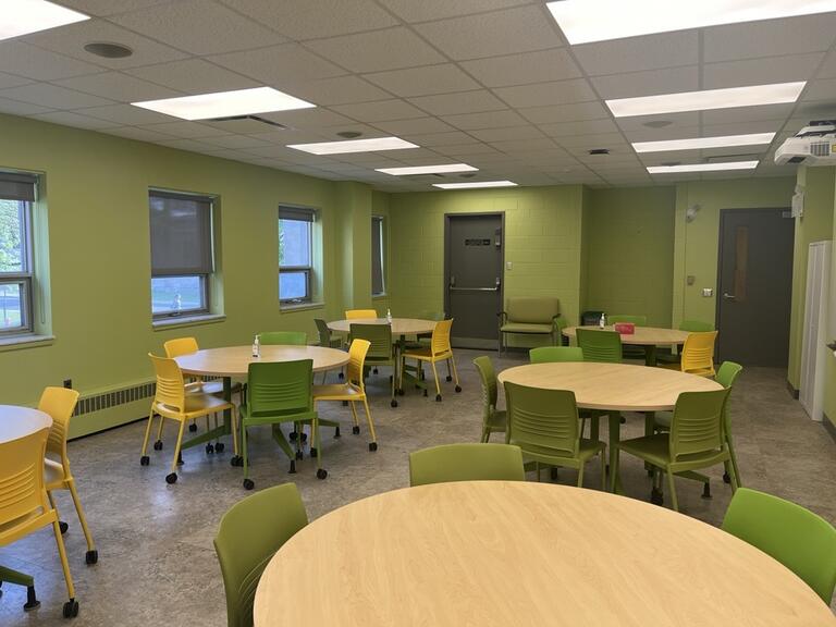 View of the classroom from the instructor’s podium. There are multiple round tables with chairs for students. Two projectors on the ceiling project onto screens on the right hand side of the room. This classroom is accessible for instructors.