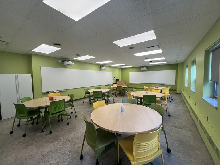 View of the classroom from a student seat near the rear of the room. In front are multiple round tables and chairs. The instructor podium is at the front of the room on the left. Digital projectors on the ceiling, projects onto screens on the front wall and left wall. This classroom is accessible for students.