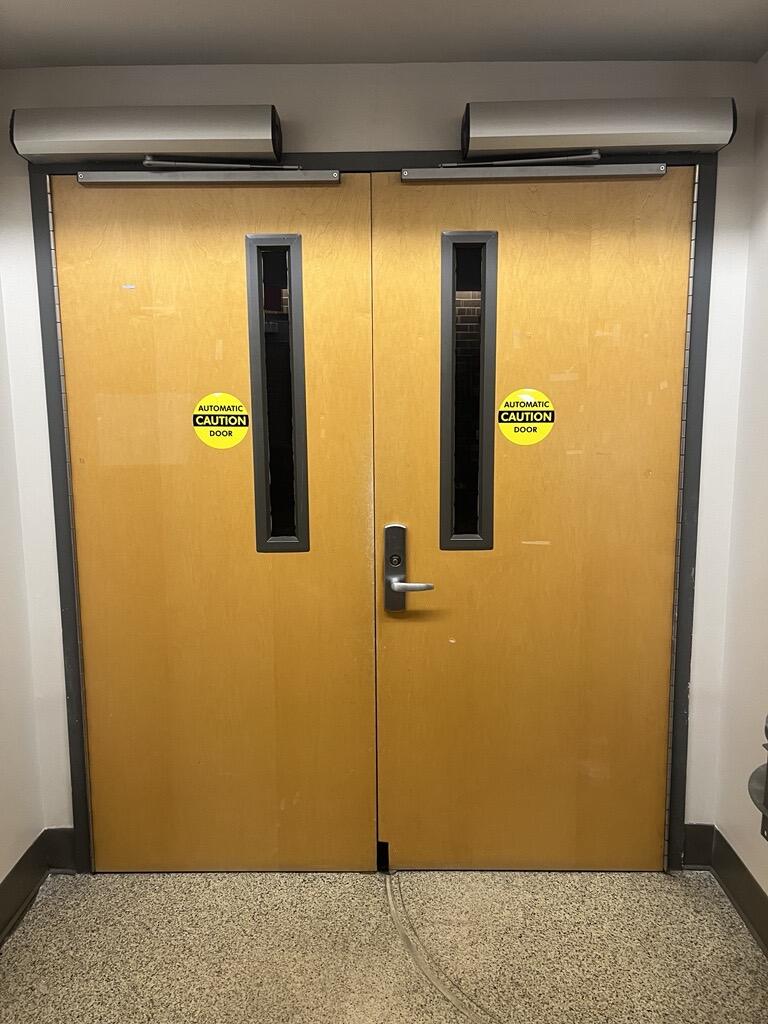 The entryway of the Auditorium in Humphrey Hall is a double door with a silver doorknob on the right door. There is an automatic door operator on the right of the doors.