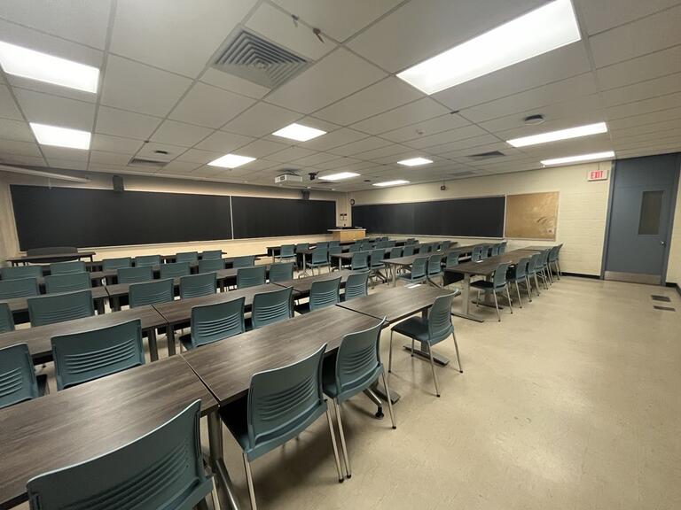 View of the classroom from a student seat near the rear of the room. In front are four empty rows of tables and chairs. The instructor podium is at the front of the room on the right. The front of the classroom has a three large blackboard panels in the middle of the front wall. A digital projector on the ceiling, projects onto a screen that extends from the ceiling. This classroom is accessible for students.