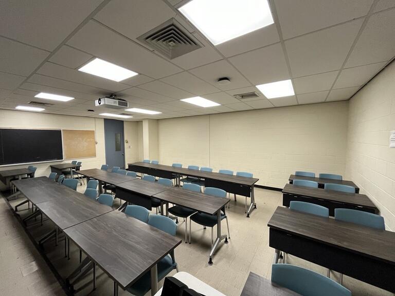 View of the classroom from the instructors podium. There are three rows of tables and chairs, with exits at the left of the room. 