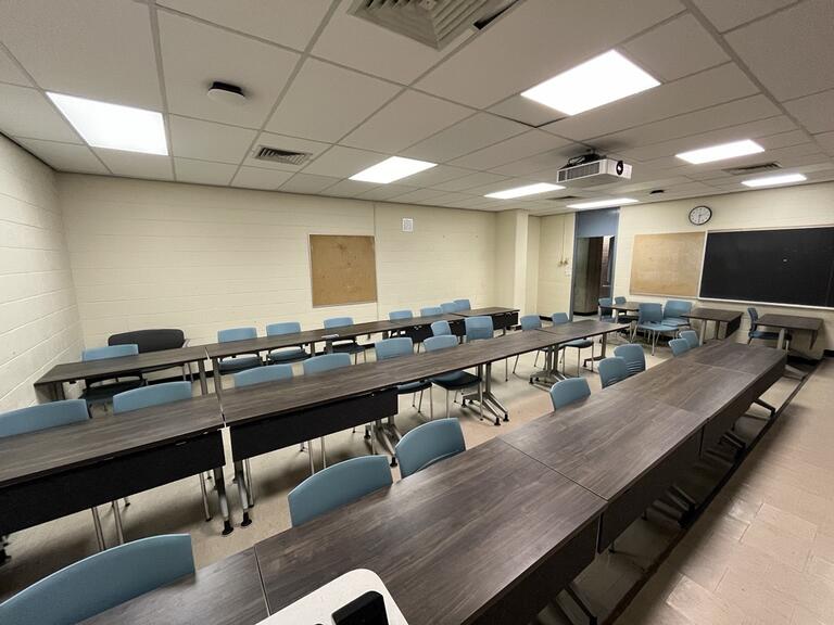 This image shows the instructors view in the room. The are three rows of desks and chairs that are both portable. At the back of the room there is a cork board on the wall. The exit is on the right side of the room.