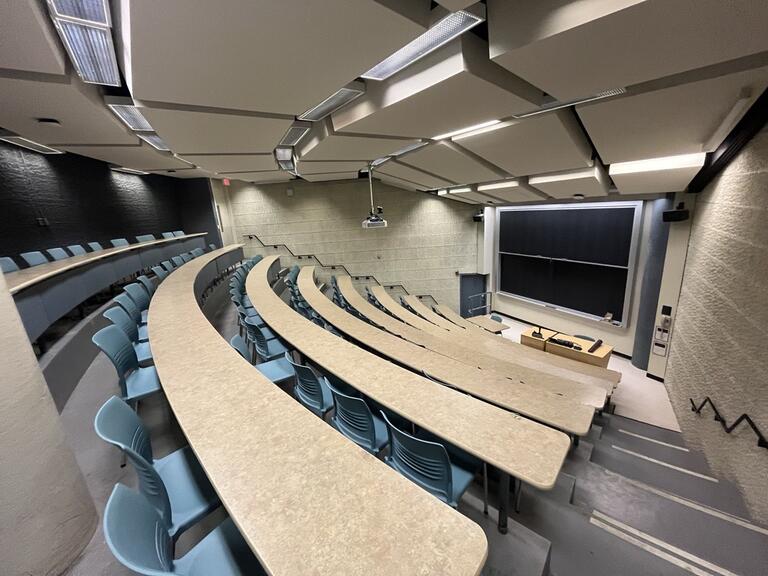 View of the classroom from a student seat near the rear of the room. In front are multiple empty rows of tables and chairs. The instructor podium is at the front of the room. The front of the classroom has a large sliding blackboard panel on the front wall. This classroom is not accessible for students.