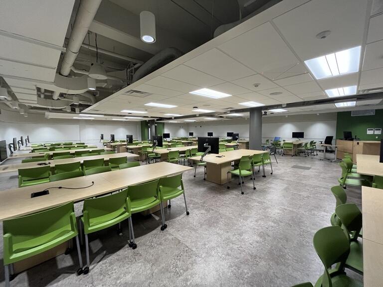 View of the classroom from a student seat. The podium is in the corner of the room and has whiteboards behind it. There is a computer at the podium. The podium is height adjustable and is accessible for instructors.