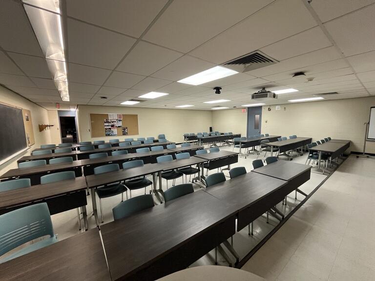 View of the classroom from the instructor’s podium. There are four rows of tables with chairs for students. A blackboard is located on the right wall. This classroom is not accessible for instructors. The podium is equipped with a document camera, lecture capture and a set of microphones.