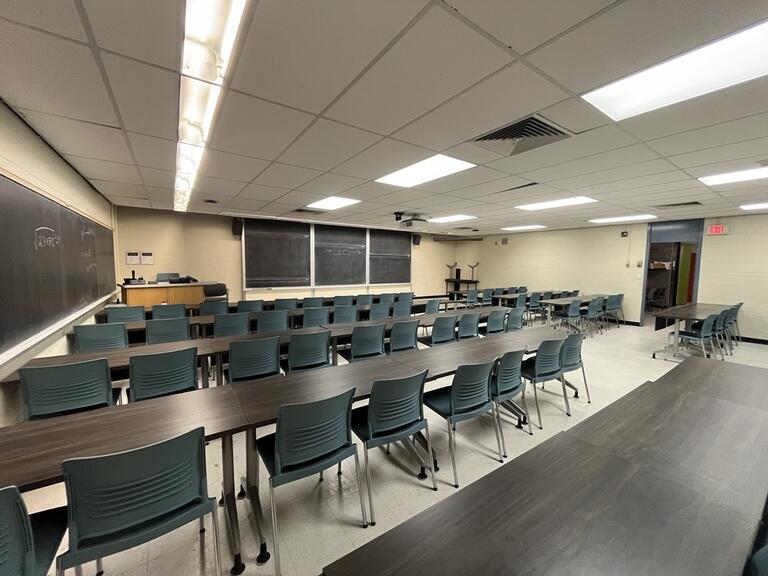 View of the classroom from a student seat near the rear of the room. In front are multiple empty rows of tables and chairs. The instructor podium is at the front of the room on the left. Three large sliding blackboard panels are located on the front wall. A projector on the ceiling projects onto a screen that extends from the ceiling. This classroom is accessible for students.