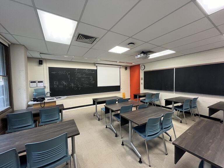 View of the classroom from a student seat near the rear of the room. In front are multiple empty rows of tables and chairs. The instructor podium is at the front of the room on the left. Two blackboard panels are located on the front wall. A projector on the ceiling projects onto a screen that extends from the ceiling. This classroom is accessible for students.