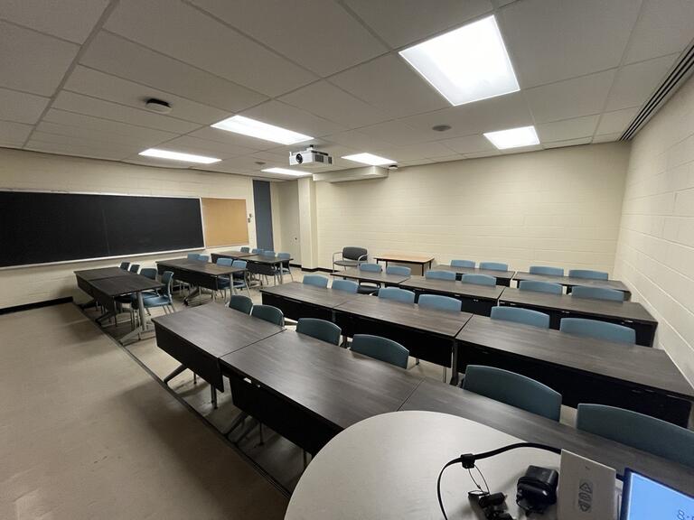 View of the classroom from the instructor’s podium. There are multiple tables with chairs for students, arranged in rows facing the front of the room. The entryway door is located on the left side of the classroom. This classroom is not accessible for instructors.