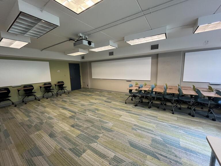 View of the classroom from the instructor’s podium. There are multiple tables and chairs for students, that can be arranged according to the instructor’s preference. Whiteboards are located on the back and right walls of the classroom. The entryway door is located at the back of the classroom, on the right. This classroom is accessible for instructors.