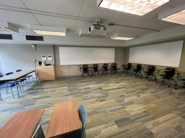 View of the classroom from a student seat near the rear of the room. In front are multiple empty chairs with tables attached. The instructor’s podium is located at the front of the room on the right. A projector on the ceiling projects onto a screen that extends from the ceiling, at the front of the room. This classroom is accessible for students.