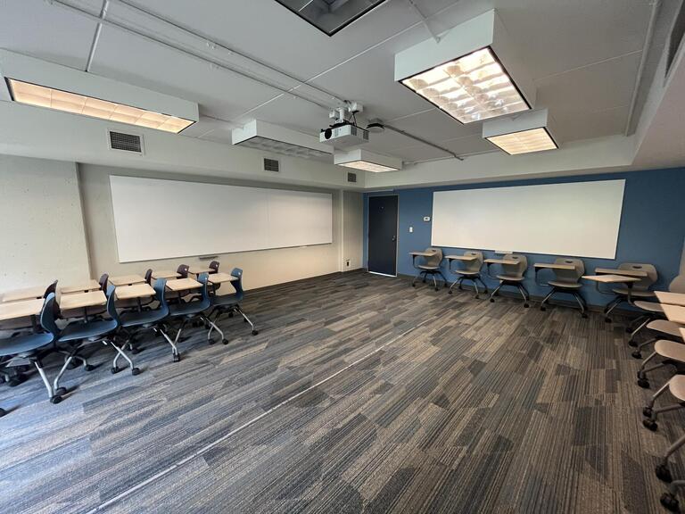 View of the classroom from the instructor’s podium. There are multiple tables with chairs attached for students, which can be arranged according to instructor’s preference. A whiteboard is located at the back of the room. The entryway door is located at the front of the room on the right. This classroom is accessible for instructors.