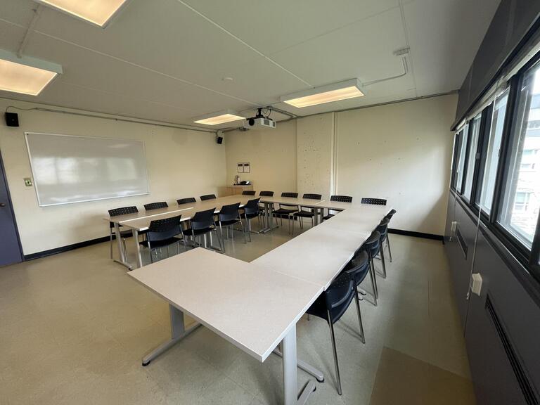 View of the classroom from a student seat near the rear of the room. In front are multiple empty tables and chairs. A whiteboard is located on the front wall. A projector on the ceiling projects onto a screen that extends from the ceiling. This classroom is accessible for students.