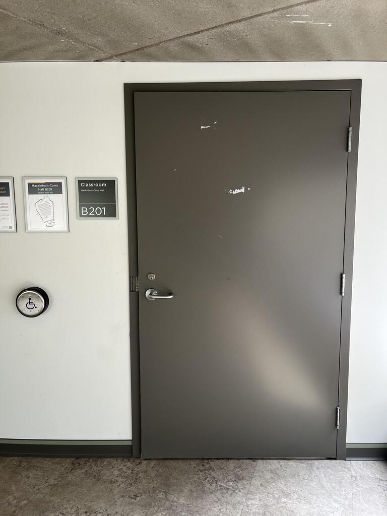 The entrance to room B201 in Mackintosh-Corry Hall is a dark grey door with a lever on the left side and an automatic operator. There is another accessible door for instructor's to access the podium, around the corner.