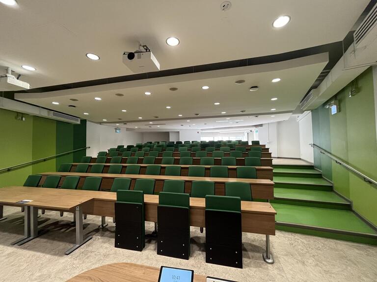 View of the classroom from the podium. The rows of seating have tiers towards the back of the room. There are stairs on either side of the room to access the back tiers. The seats are attached to the desks and there is no power at the tables. At the front of the room are two wheelchairs accessible seats with chairs pulled up to the desks.