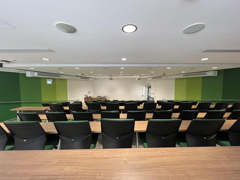 View of the classroom from a student seat near the rear of the room. The podium is at the front of the room on the left. When presenting, the image is projected on the wall to the right of the instructor’s podium.