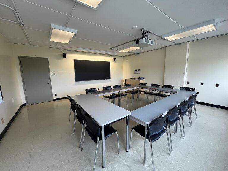 View of the classroom from a student seat near the rear of the room. In front are multiple empty tables and chairs. A blackboard is located on the front wall. A projector on the ceiling projects onto a screen that extends from the ceiling. The entryway door is located at the front of the room on the left. This classroom is accessible for students.