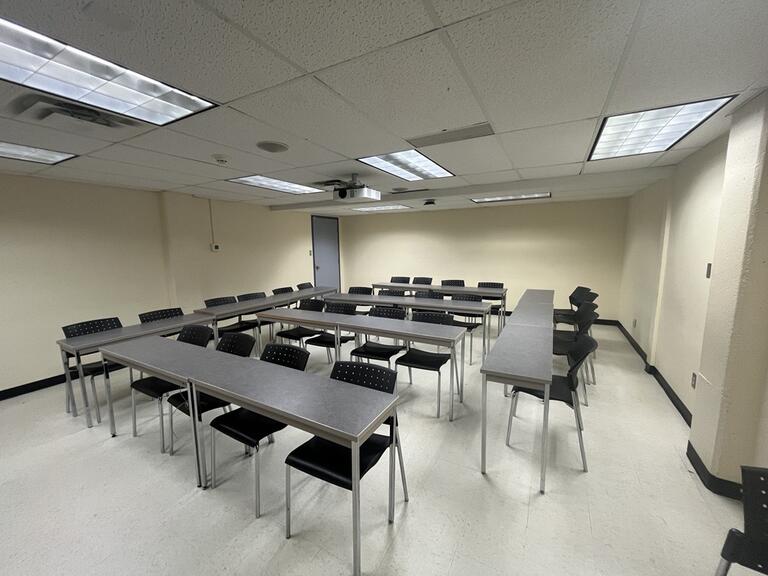 View of the classroom from the instructor’s podium. There are tables and chairs for students, arranged in a rectangle. The entryway door is located at the back of the classroom, on the left. This classroom is accessible for instructors.