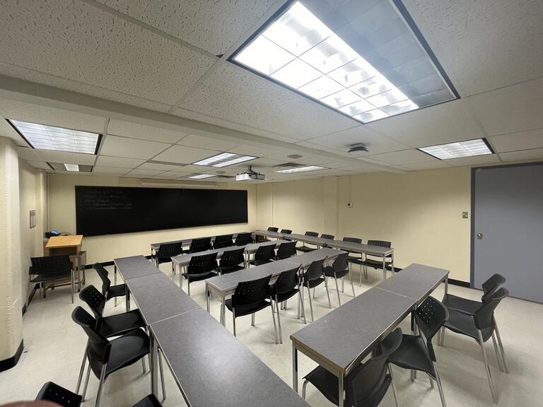 View of the classroom from a student seat near the rear of the room. In front are multiple empty tables and chairs. The instructor’s podium is at the front of the room on the left. A long blackboard is located on the front wall. A projector on the ceiling projects onto a screen that extends from the ceiling. This classroom is accessible for students.