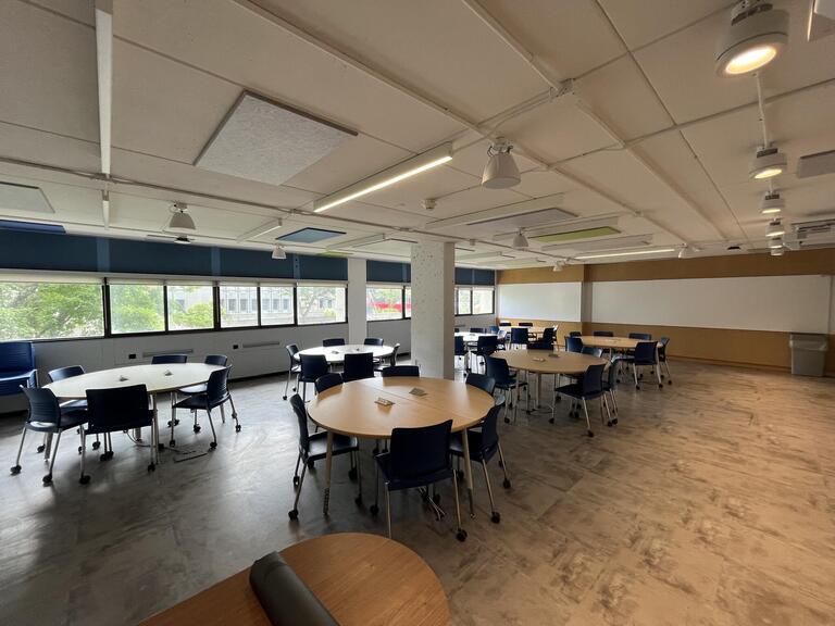 View of the classroom from the instructor's podium. In front are several round tables, each with 4 chairs. Each table has 4 total outlets. This classroom is accessible for instructors.