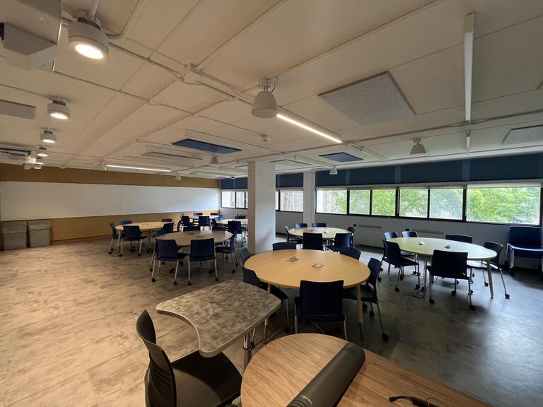 View of the classroom from the instructor's podium. In front are several round tables, each with 4 chairs. Each table has 4 total outlets. This classroom is accessible for instructors.