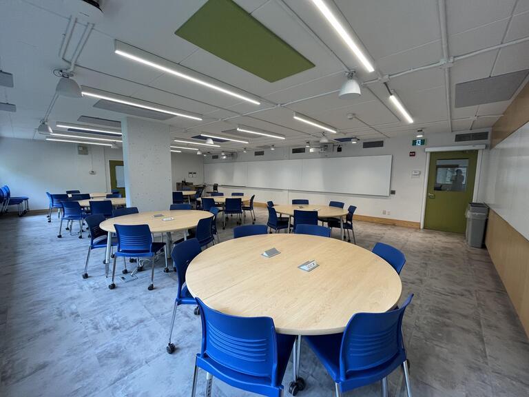 View of the classroom from the student's perspective. In front are several round tables with 4 chairs each. Each table has 4 outlets. The projector's on the wall project onto the whiteboards around the room. This classroom is accessible for students.