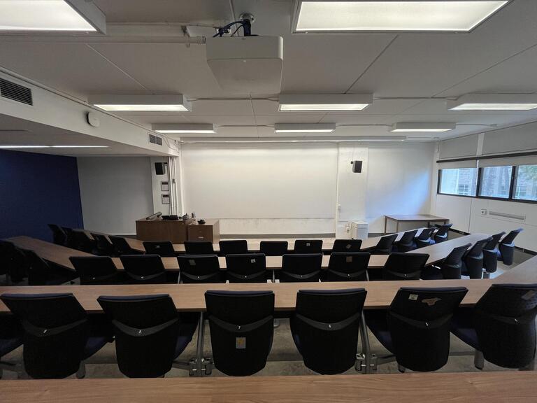 View of the classroom from a student seat near the rear of the room. In front are rows of wood tables and blue chairs. The instructor’s podium is at the front of the room on the left. The front wall of the classroom is covered in a whiteboard surface. A digital projector on the ceiling projects onto the whiteboard surface at the front of the classroom.