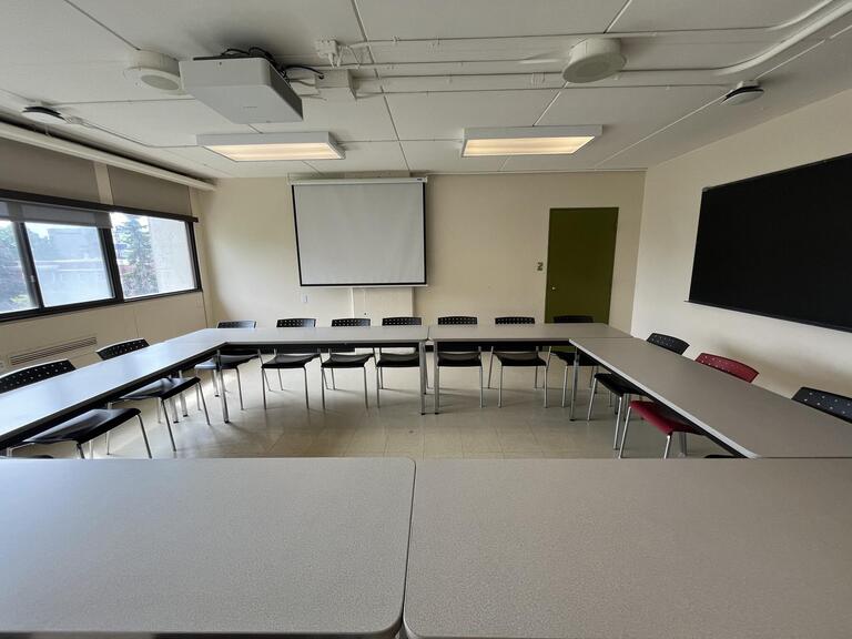 View of the classroom from the front of the classroom. There are multiple tables and chairs for students, arranged in a square in the centre of the room. This classroom is accessible for instructors.