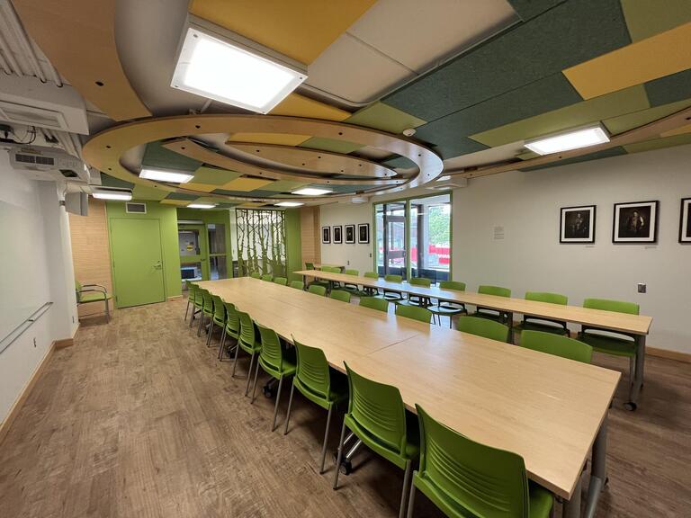 View of the classroom from the front of the classroom. There are multiple tables and chairs for students, arranged in a straight line in the centre of the classroom. This classroom is accessible for instructors.